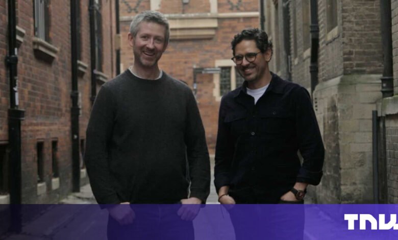 Two men stand side-by-side in a brick alleyway, smiling at the camera.