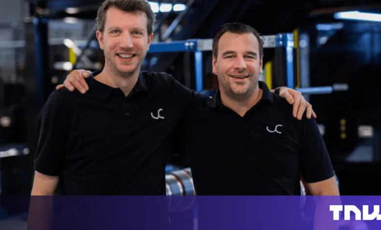 Two smiling men in black shirts stand with arms around each other in a factory setting.