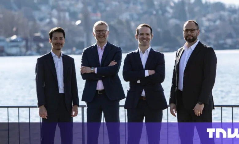 Four men in suits stand outdoors near a lake.