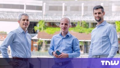 Three men in light blue shirts stand outdoors, smiling at the camera.