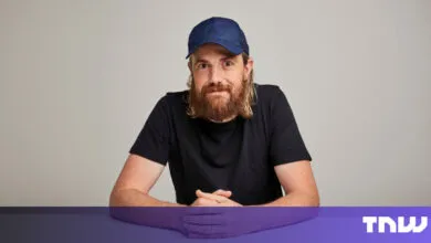 Man with beard and cap sits at table, hands clasped.