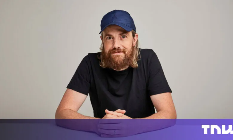 Man with beard and cap sits at table, hands clasped.