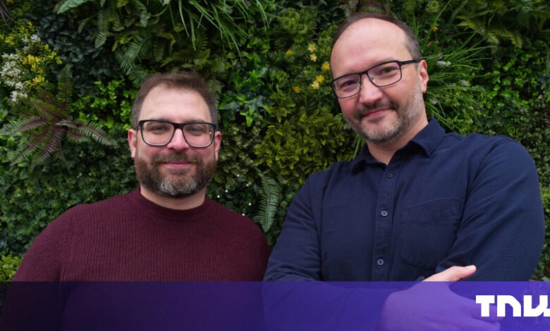 Two men smiling in front of a lush green wall.