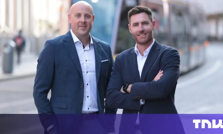 Two men in suits stand outdoors, smiling confidently.