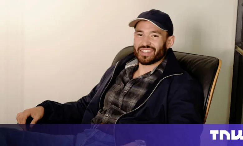 Man with beard smiles, wearing cap and jacket in chair.