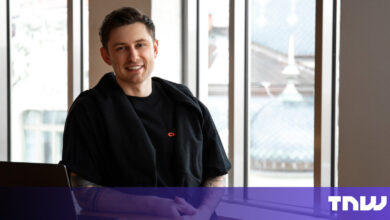 Smiling man in black shirt sits at desk with laptop.