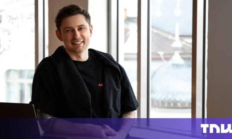 Smiling man in black shirt sits at desk with laptop.