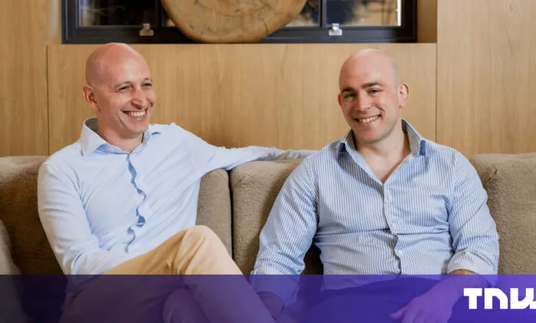 Two smiling men sit on a couch in a modern, light-filled room.
