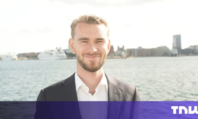 Smiling man in suit stands by water with city skyline in background.