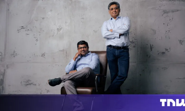 Two men pose in front of a textured gray wall, one seated in a leather chair.