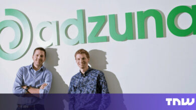 Two men stand in front of the Adzuna logo, smiling at the camera.