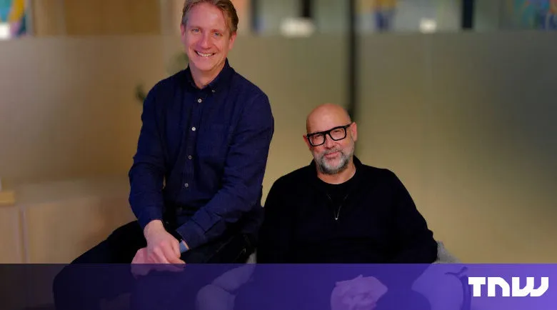 Two men pose indoors, one seated, one perched, smiling warmly.