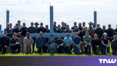 Group poses with large drone in grassy field under a bright sky.