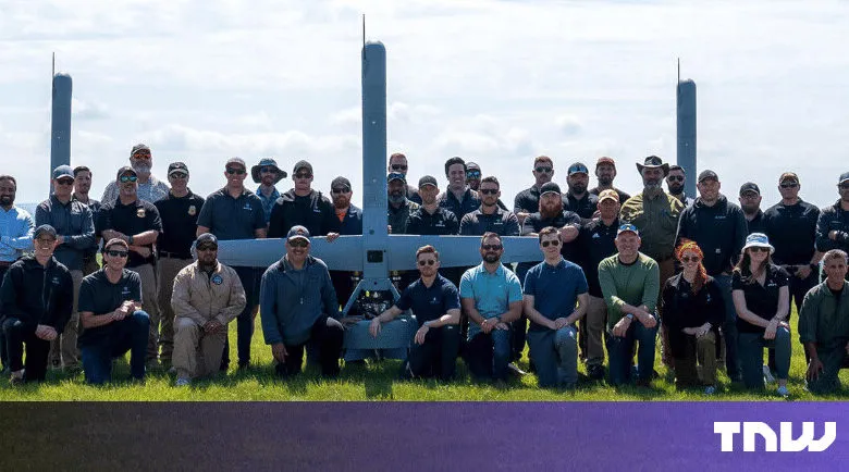 Group poses with large drone in grassy field under a bright sky.