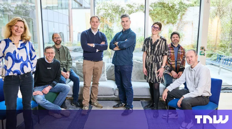 Group of eight people posing indoors with large windows and natural light.