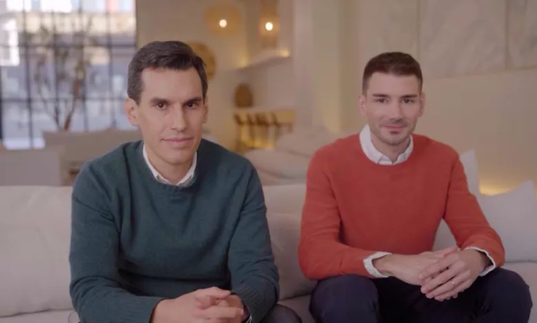 Two men sit side-by-side on a couch, looking directly at the camera.