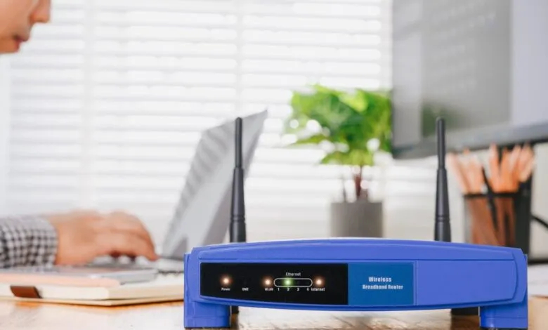 Blue wireless router on desk with person working on laptop in background.