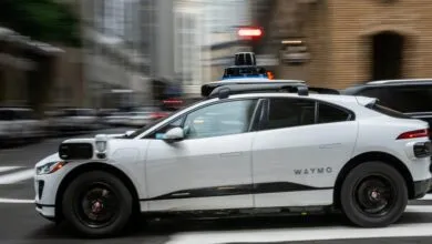 White Waymo self-driving car driving on city street with sensor equipment.