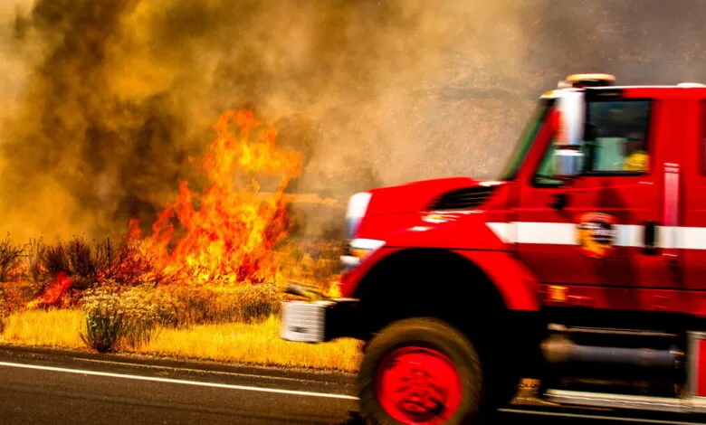 Red fire truck speeding past a brush fire with flames and smoke.