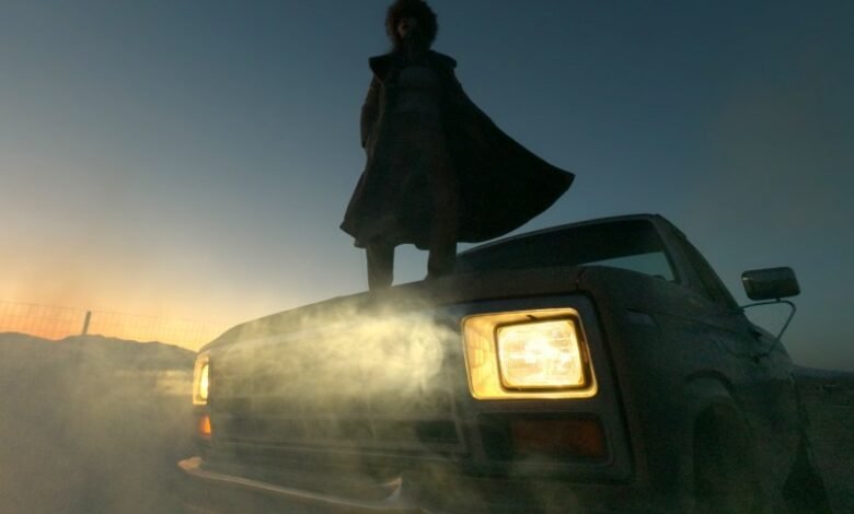 Person in coat stands atop vintage truck with headlights on at dusk.