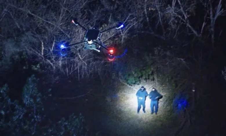 Drone with blue and red lights hovers over two figures in a dark, wooded area.
