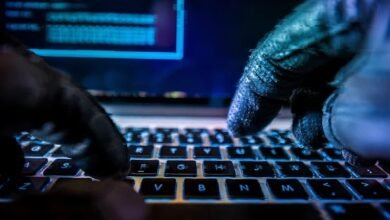 Gloved hands type on a laptop keyboard in a dark, blue-lit environment.