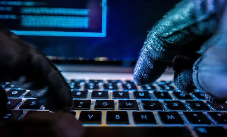Gloved hands type on a laptop keyboard in a dark, blue-lit environment.