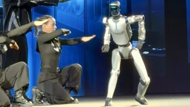 Dancers in black perform alongside a humanoid robot on a stage.