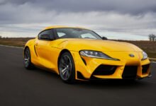 A bright yellow Toyota Supra driving on a paved road under a cloudy sky.