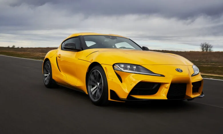 A bright yellow Toyota Supra driving on a paved road under a cloudy sky.