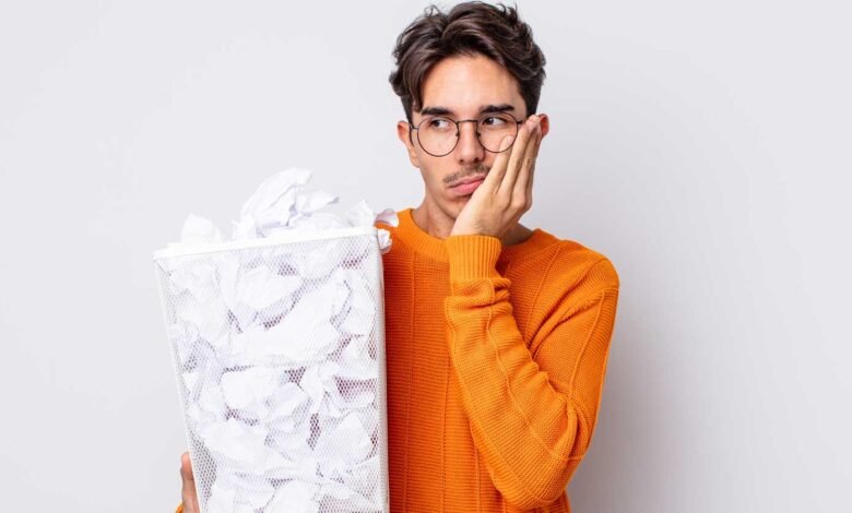Man with glasses looking bored holding a trash can full of crumpled paper.