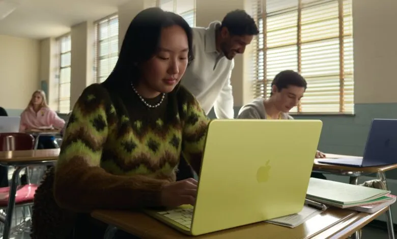 Student uses a yellow MacBook in a classroom setting with teacher nearby.