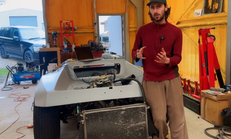 Man stands next to a partially assembled car in a garage setting.