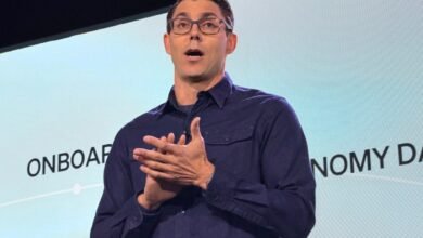 RJ Scaringe, Rivian CEO, presents onstage with hands clasped, wearing glasses and a blue shirt.