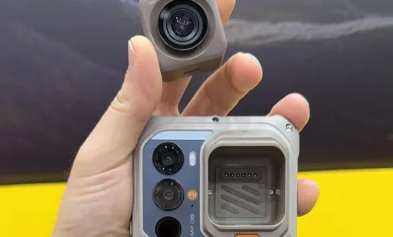 Hand holding modular camera attachment above rugged smartphone.
