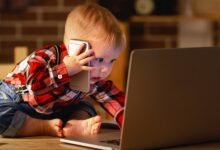 Baby in plaid shirt uses laptop and smartphone, looking intently at the screen.