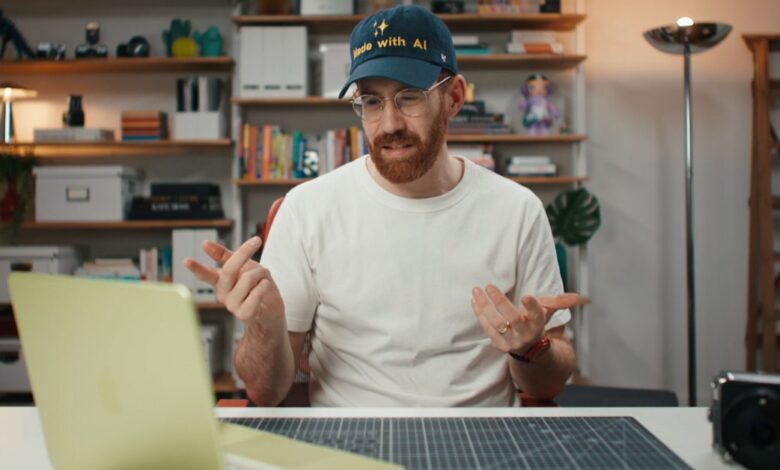 Man with beard and glasses wearing 'Made with AI' hat gestures while looking at laptop.