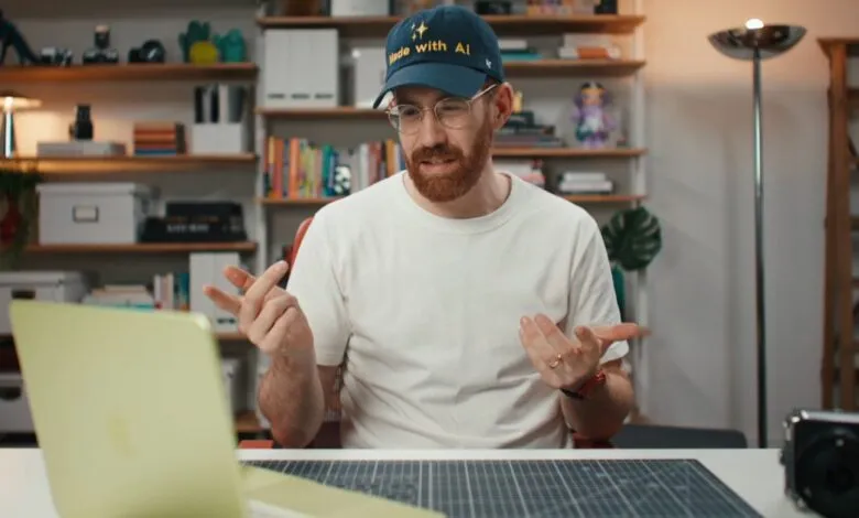 Man with beard and glasses wearing 'Made with AI' hat gestures while looking at laptop.