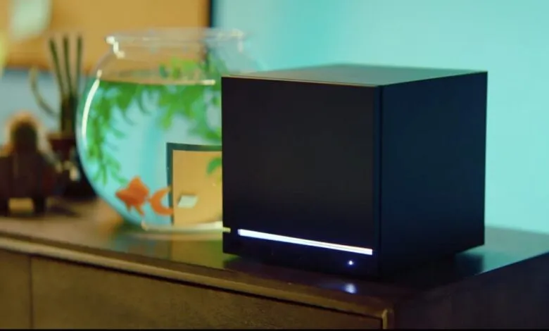 Black cube device sits on a wooden surface next to a fishbowl.