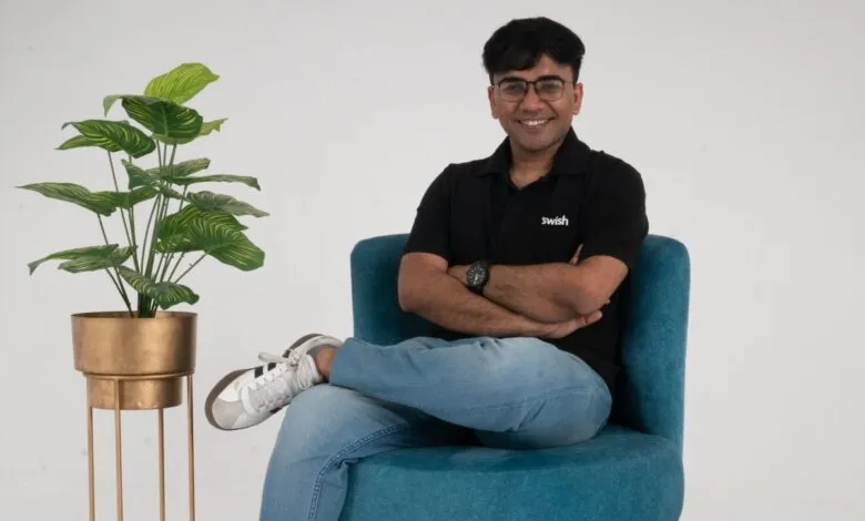 Man in black shirt with 'swish' logo sitting in blue chair, smiling.