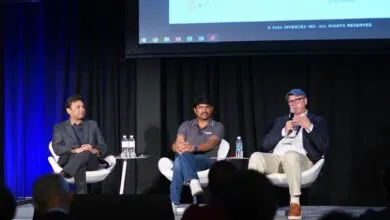 Three men on stage during a panel discussion, one speaking into a microphone.