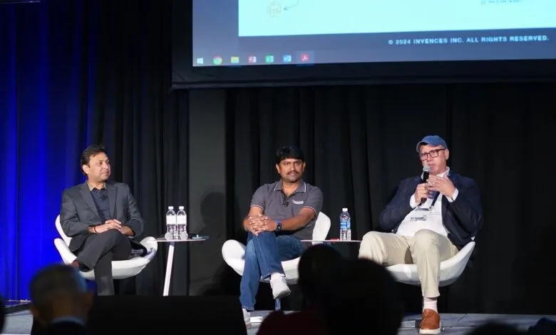 Three men on stage during a panel discussion, one speaking into a microphone.