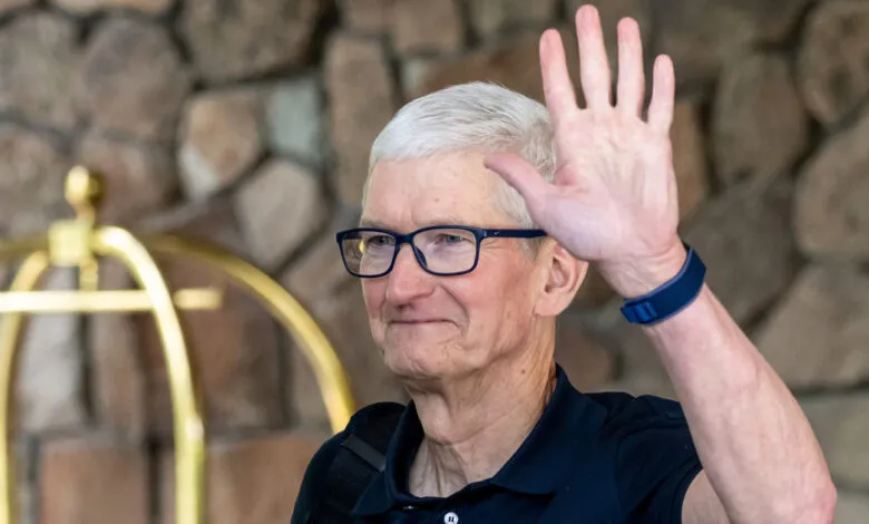 Tim Cook waves with a friendly smile, wearing glasses and a dark shirt.