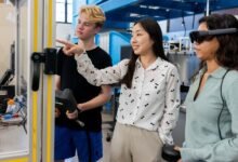 Three students in a lab setting, one pointing at a robotic arm, another wearing a VR headset.