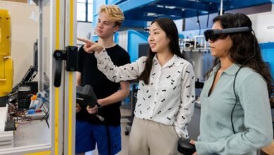 Three students in a lab setting, one pointing at a robotic arm, another wearing a VR headset.