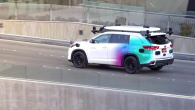 Zoox self-driving car with sensor array on road, white with colorful gradient.