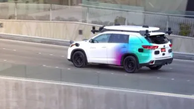 Zoox self-driving car with sensor array on road, white with colorful gradient.