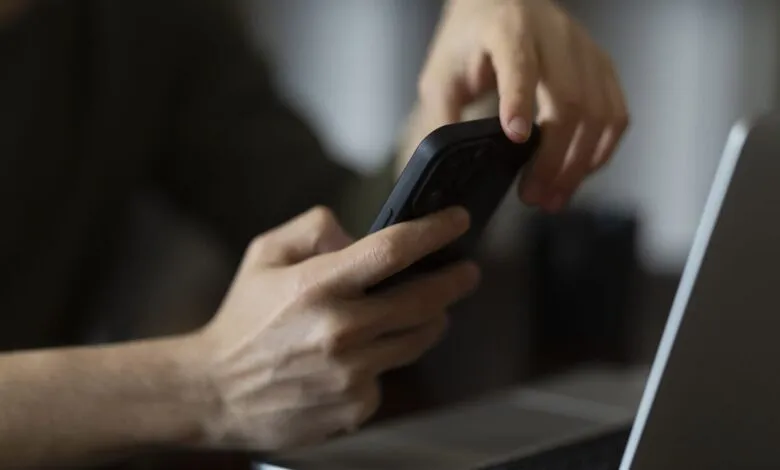 Hands holding smartphone next to laptop, blurred background.