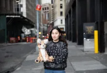 Woman holds deer plushie on city street, buildings in background.