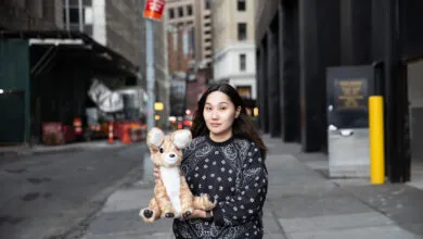 Woman holds deer plushie on city street, buildings in background.